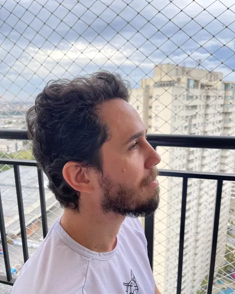 Antes do corte: cliente de perfil com cabelo ondulado e barba aparada, na varanda com tela de proteção