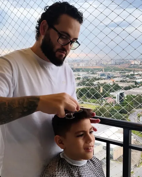 Corte de cabelo infantil na varanda Barbeiro de camiseta branca cortando o cabelo de um menino na varanda, com a cidade ao fundo