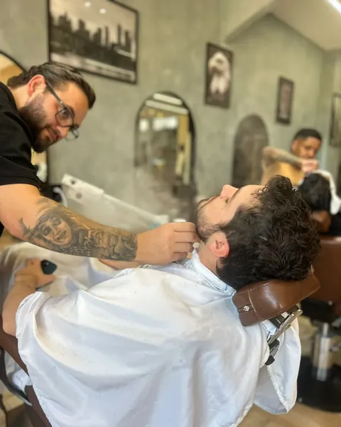 Acabamento da barba Barbeiro com tatuagem no braço ajustando a barba de um cliente de cabelo cacheado, de capa branca