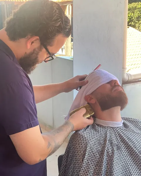 Barba com toalha e máquina dourada Barbeiro de óculos aparando a barba de um cliente com toalha sobre o rosto, com máquina dourada, na varanda