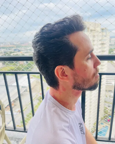 Corte com topete e barba curta Cliente de perfil com topete volumoso e barba curta, na varanda com céu e prédios ao fundo