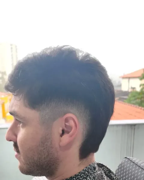Degradê alto com topete Cliente de perfil com degradê alto e topete, de capa estampada, com telhados ao fundo