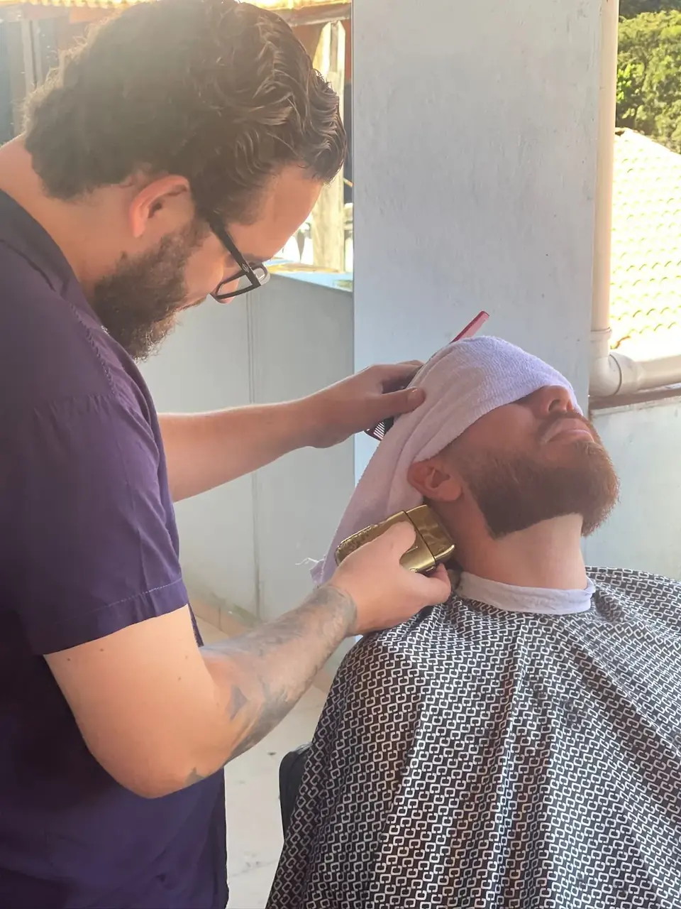 Barba feita pelo Thiego Faria Barbeiro de óculos aparando a barba de um cliente com toalha sobre o rosto e máquina dourada, na varanda