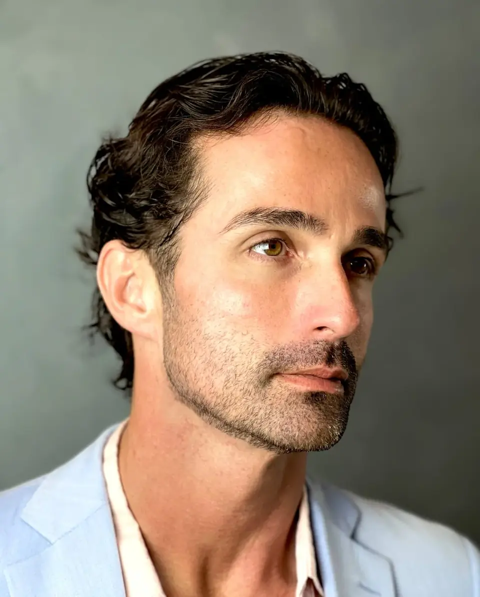 Visagismo com o Thiego Faria Retrato de homem de cabelo escuro ondulado e barba por fazer, de blazer azul-claro, em fundo cinza